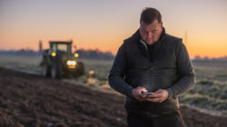 Man with short brown hair looking down while using his smartphone,standing on an agricultural field in the evening,tractor with lights in the background