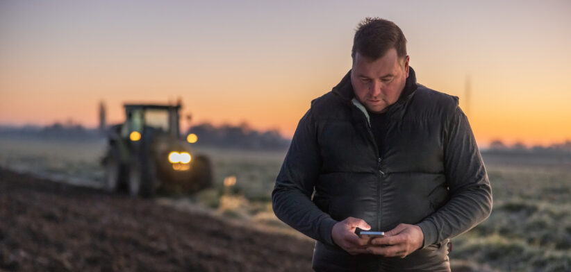 Man with short brown hair looking down while using his smartphone,standing on an agricultural field in the evening,tractor with lights in the background