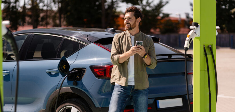 Man charging electric car and using smartphone at charging station