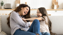 Overjoyed laughing young single mom little daughter tickling on couch