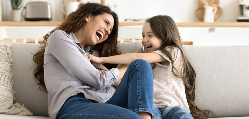 Overjoyed laughing young single mom little daughter tickling on couch